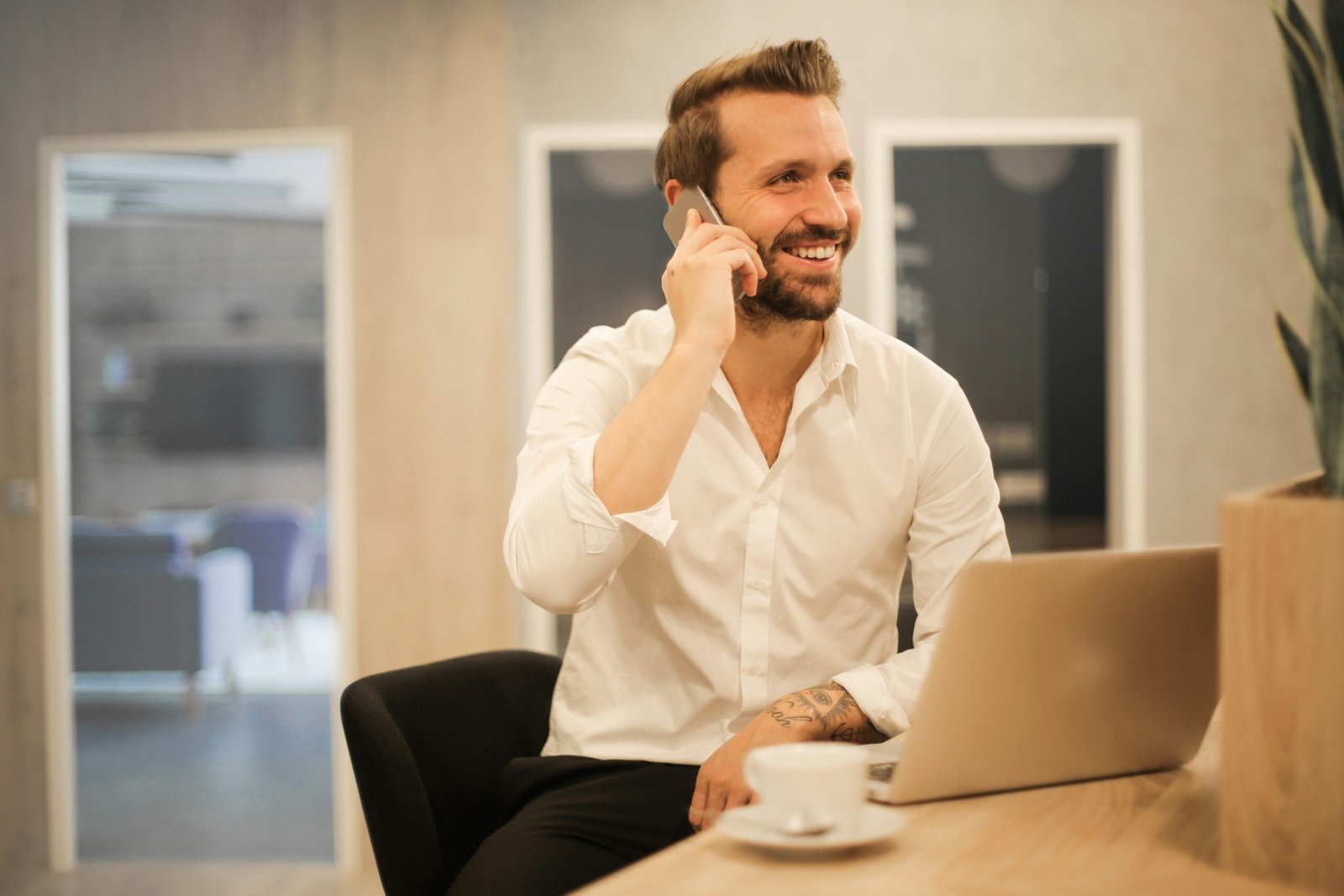 Entrepreneur Working Remotely in a Modern Cafe A smiling man with a beard and a white button-down shirt is talking on a smartphone while sitting at a wooden table with an open laptop. A white coffee cup is in the foreground, and the background shows a brightly lit, modern office or cafe interior.