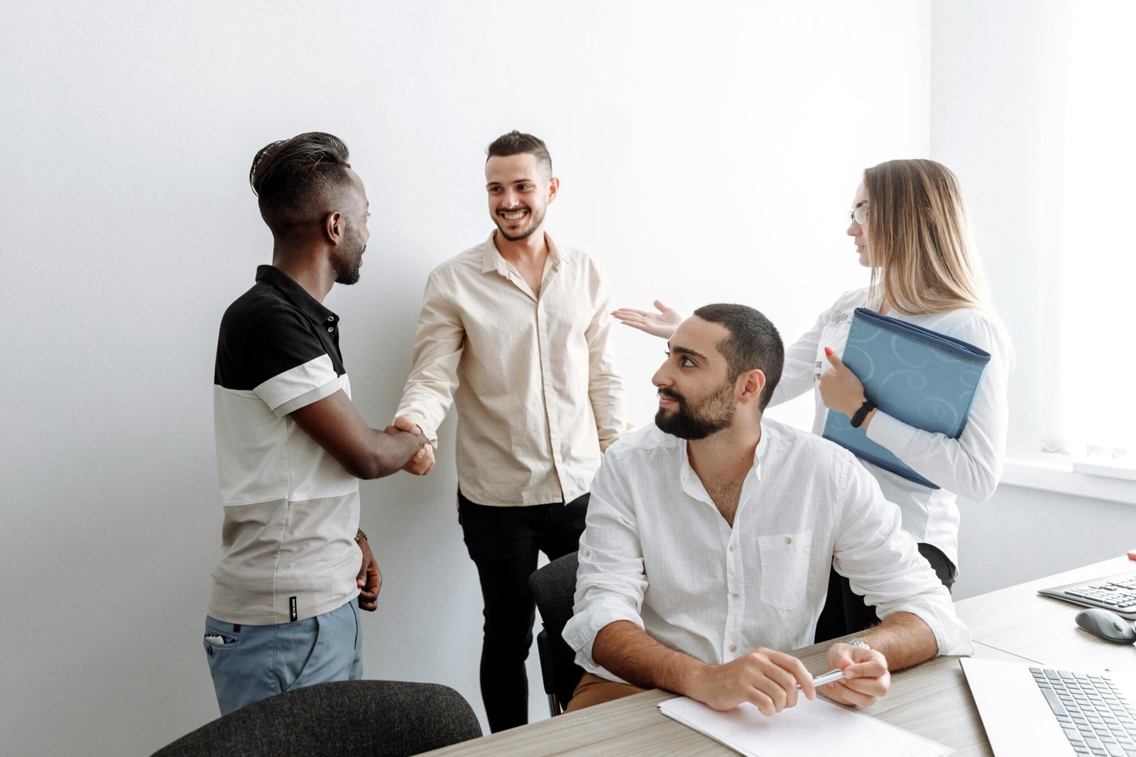A group of colleagues sitting around a long wooden table in a brightly lit, modern office kitchen. They are engaged in conversation, with some facing away from the camera. The background features open shelving, industrial-style pendant lights, and a window with blinds.