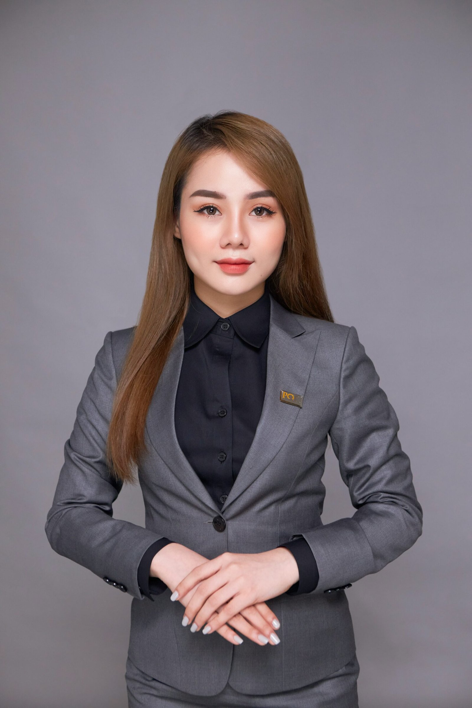 A young woman with long light-brown hair wearing a tailored gray blazer and matching trousers over a black button-down shirt. She is standing with her hands crossed in front of a solid gray background.