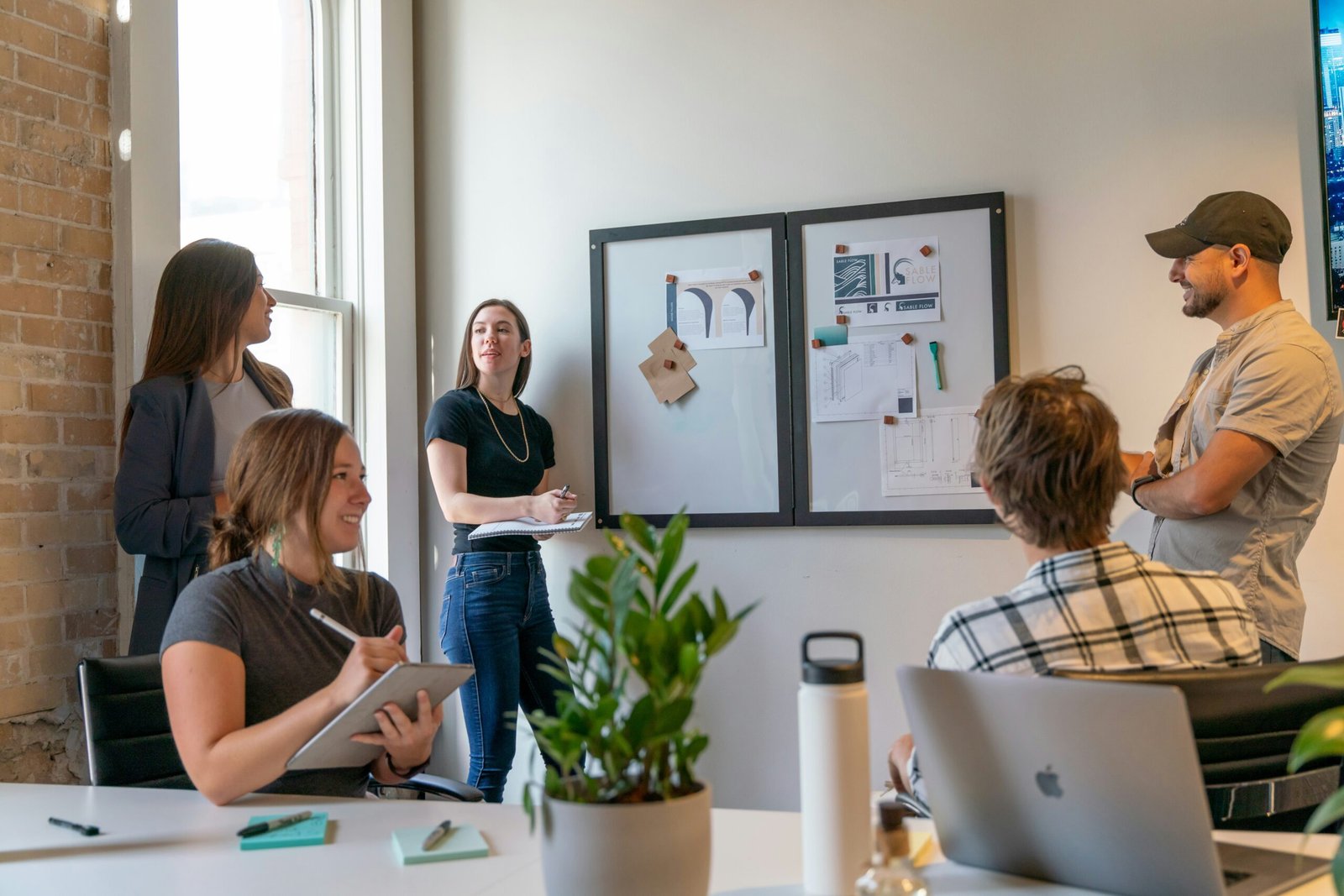 A diverse group of young professionals in a bright, modern office collaborating around a whiteboard. One woman takes notes on a tablet, another stands with a notebook, and a man in a baseball cap gestures toward design sketches pinned to the board.