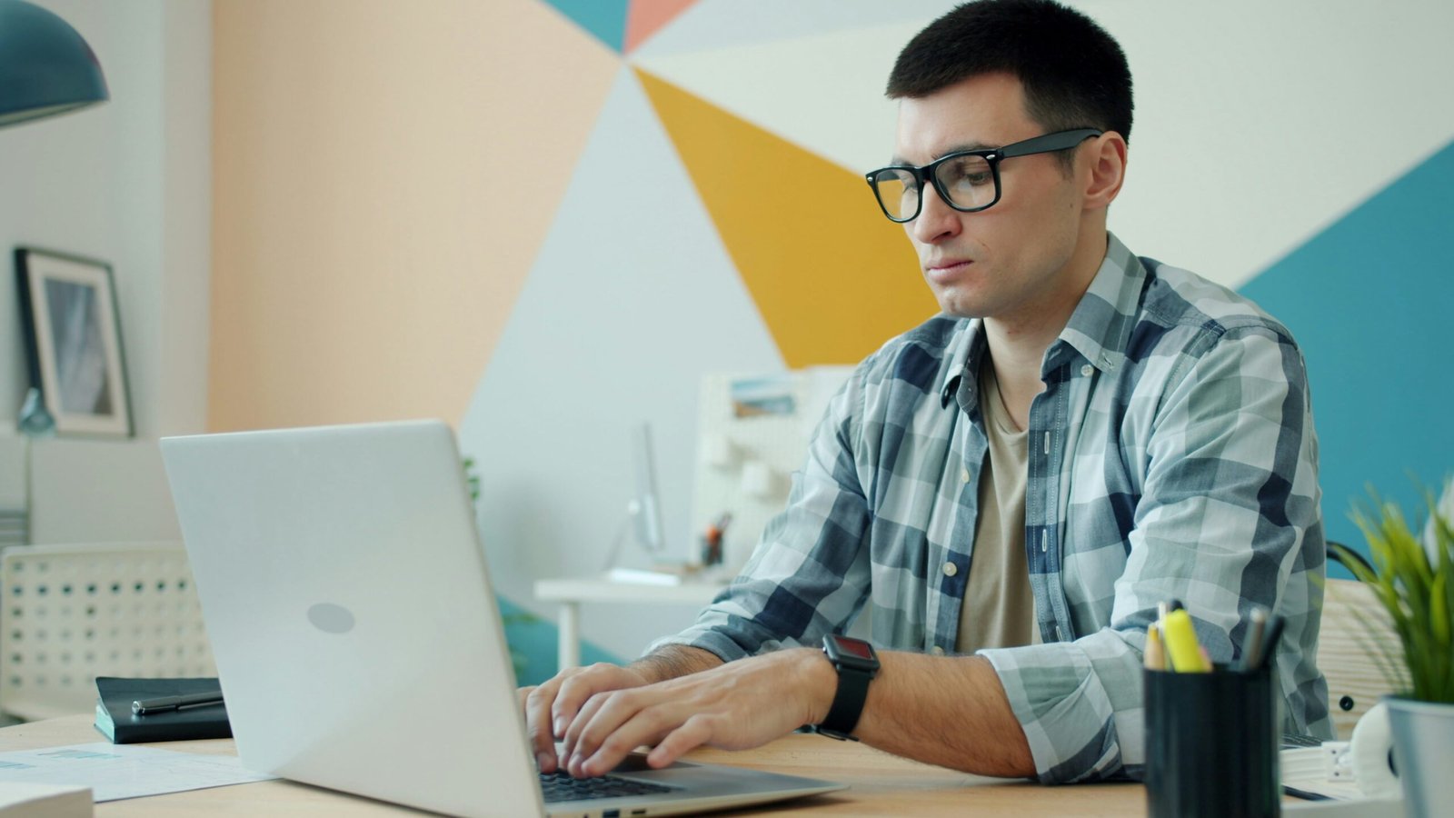 Focused Professional Working on a Laptop A man with glasses and a plaid shirt typing on a silver laptop at a wooden desk. He is in a modern office with a colorful geometric mural in the background.
